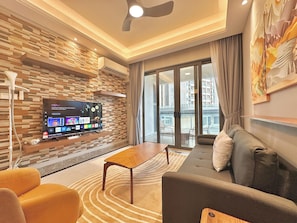 A modern living room with a flat-screen TV mounted on a textured wall, a sofa, a coffee table, and a view of the outside balcony. - R&F Princess Cove Phase 2 by Stayrene (Johor Bahru)