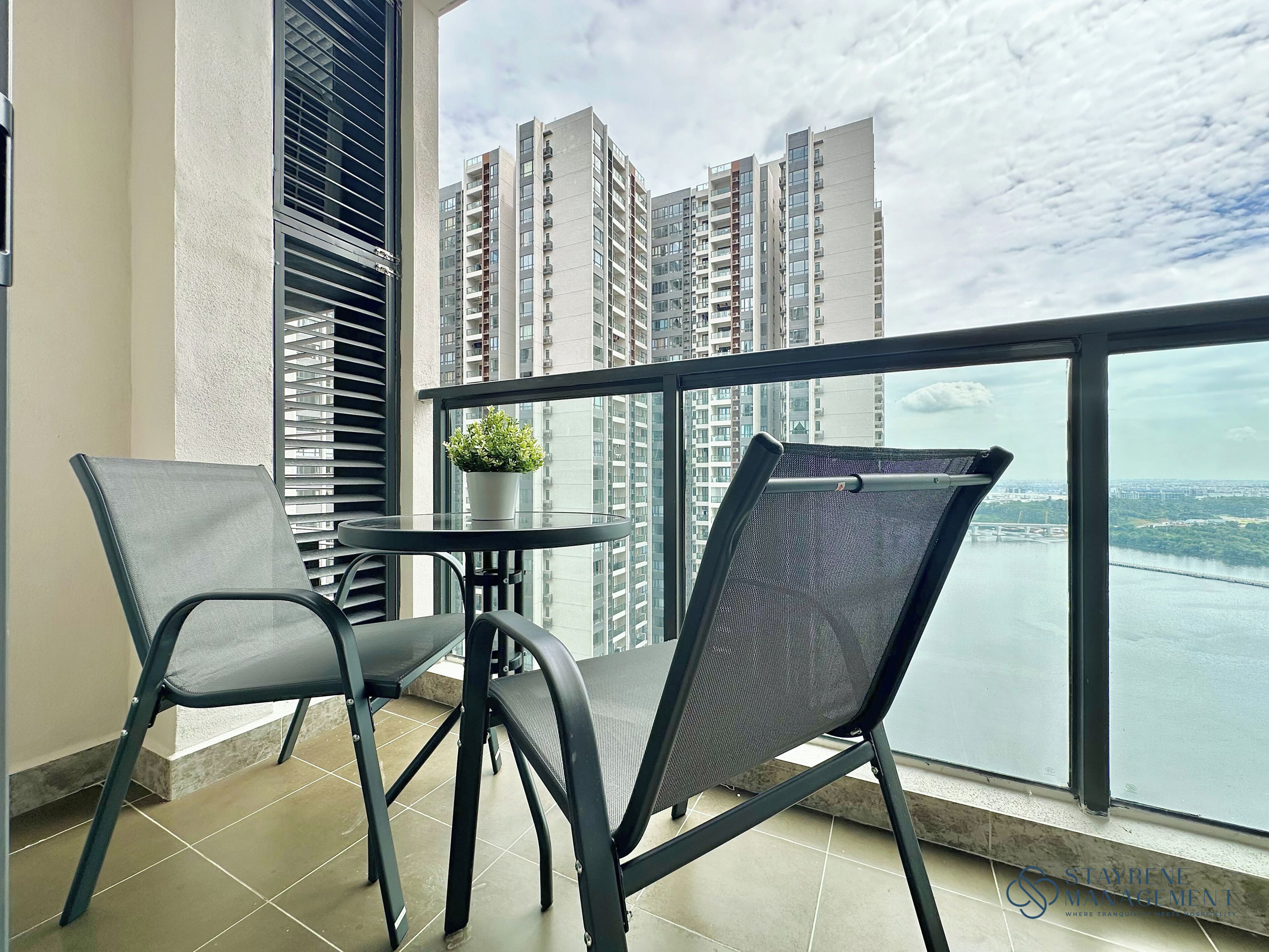 A balcony with two chairs, a small table, and a potted plant, overlooking a river and high-rise buildings.