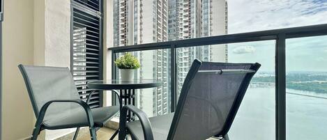 A balcony with two chairs, a small table, and a potted plant, overlooking a river and high-rise buildings.