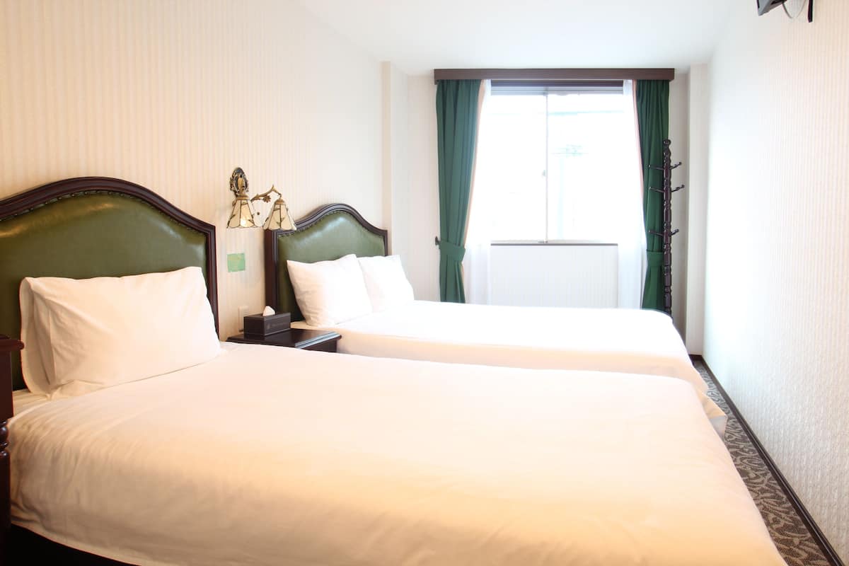 Habitación de hotel con dos camas, una mesita de noche, una lámpara y una ventana con cortinas.