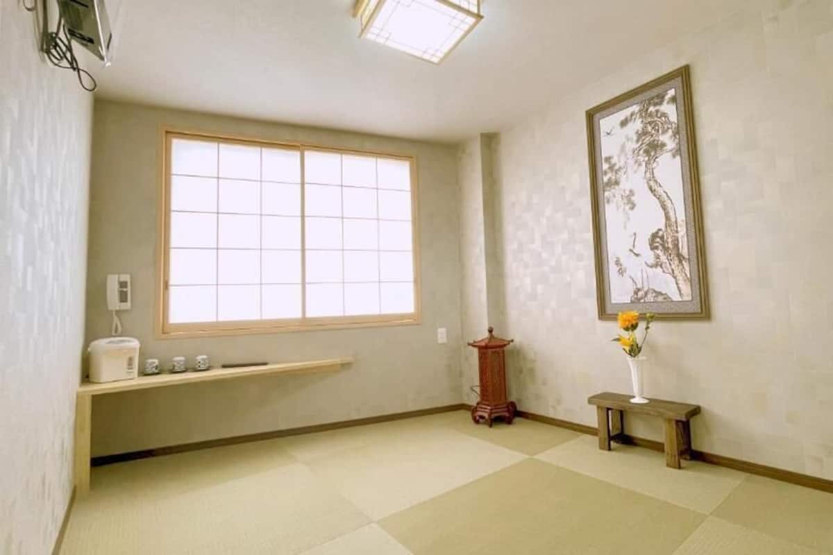 Una habitación con un ventanal amplio, un banco de madera, una mesita con un florero y una obra de arte japonesa tradicional en la pared.