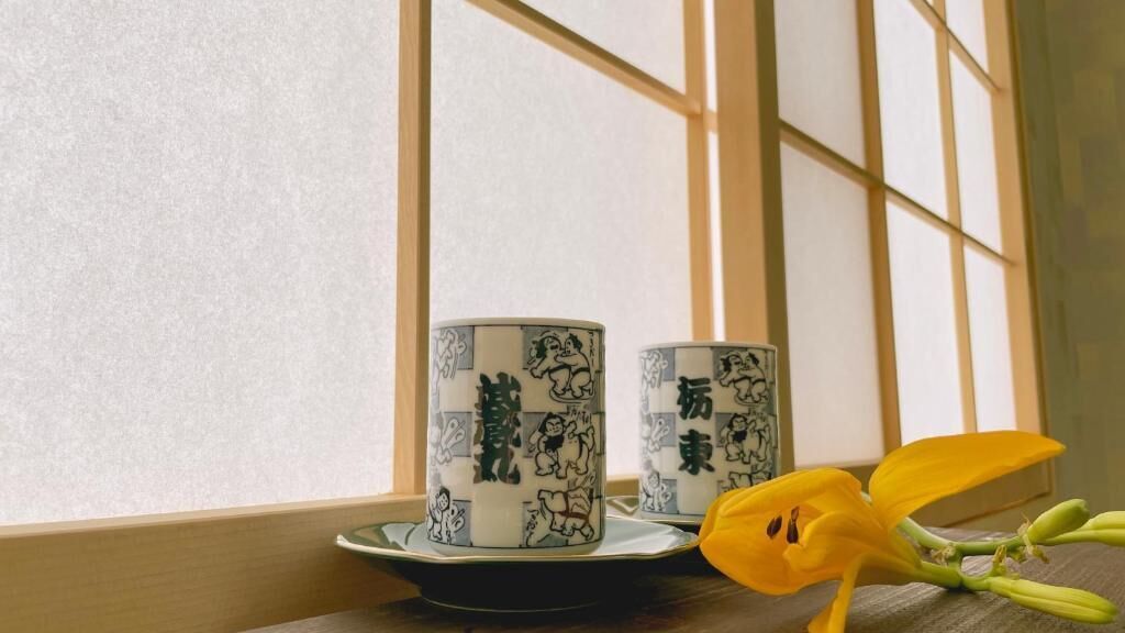 Deux tasses en céramique ornées d’illustrations noires, posées sur un plateau, sur une table près d’une fenêtre aux cadres en bois.