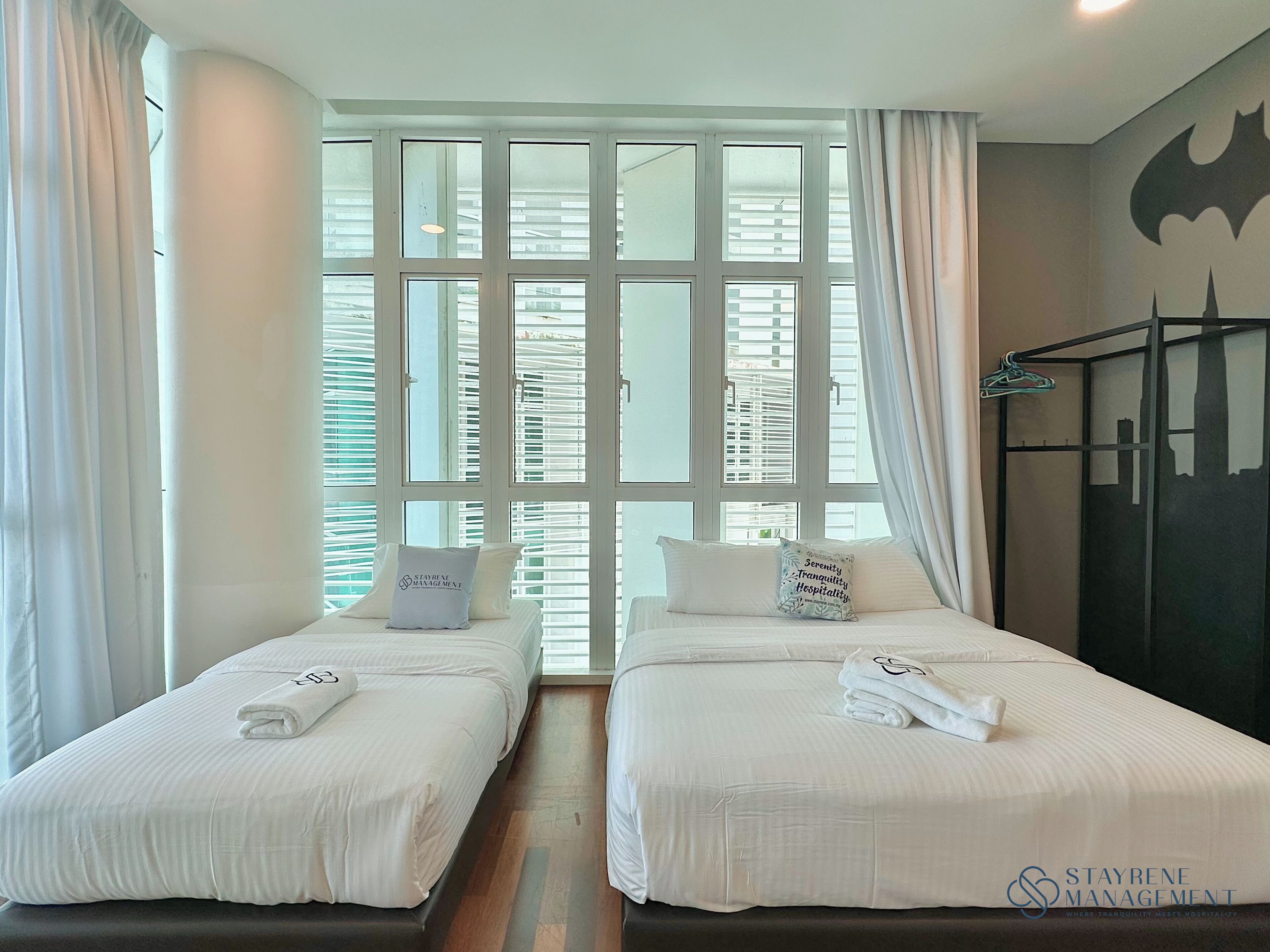 A hotel room with two beds, a large window with blinds, a coat rack, and a bat-shaped wall decoration.