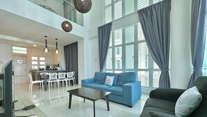 A spacious living area with a sofa, a coffee table, and a TV. There is a dining area with chairs and a table. The room has large windows with curtains. - 1 Medini Residence Neo by Stayrene (Iskandar Puteri)