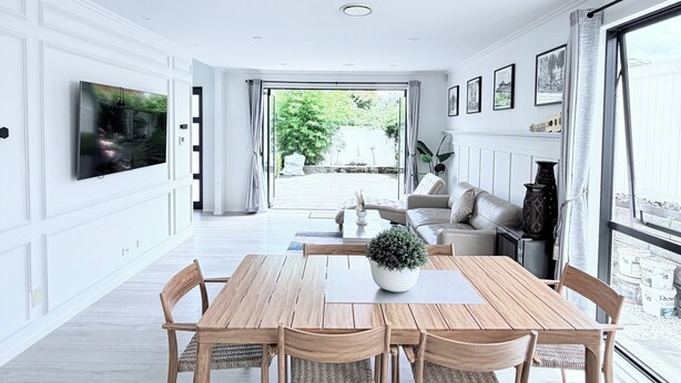 Un vaste salon doté d’un grand téléviseur, un coin repas avec une table et des chaises en bois, et une vue sur l’extérieur grâce à de grandes fenêtres.