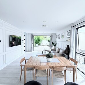 A spacious living room with a large TV, a dining area with a wooden table and chairs, and a view of the outdoors through large windows.
