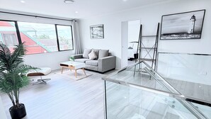 Un salon moderne avec un canapé, une table basse et une plante en pot. Un escalier avec des rampes en verre mène à un niveau supérieur.