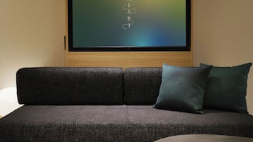 A modern living room with a dark grey sofa, a teal pillow, and a large flat-screen TV mounted on a wooden wall.