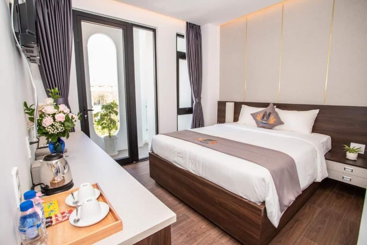 A modern hotel room with a large bed, a bedside table with a tray, a coffee maker, and a view of the outdoors.