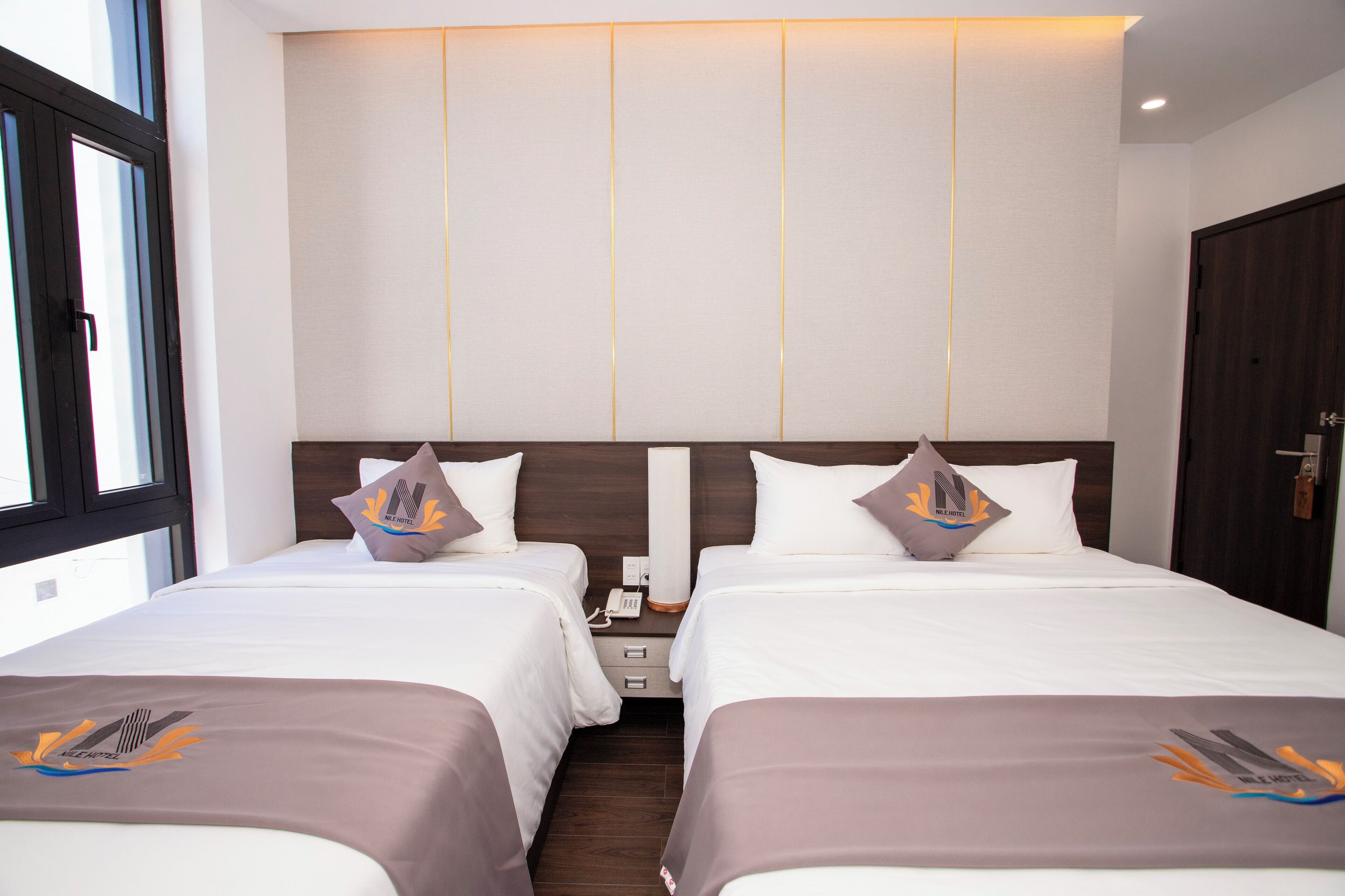 A hotel room with two beds, each with a pillow featuring a logo, a nightstand with a phone, and a window providing natural light.
