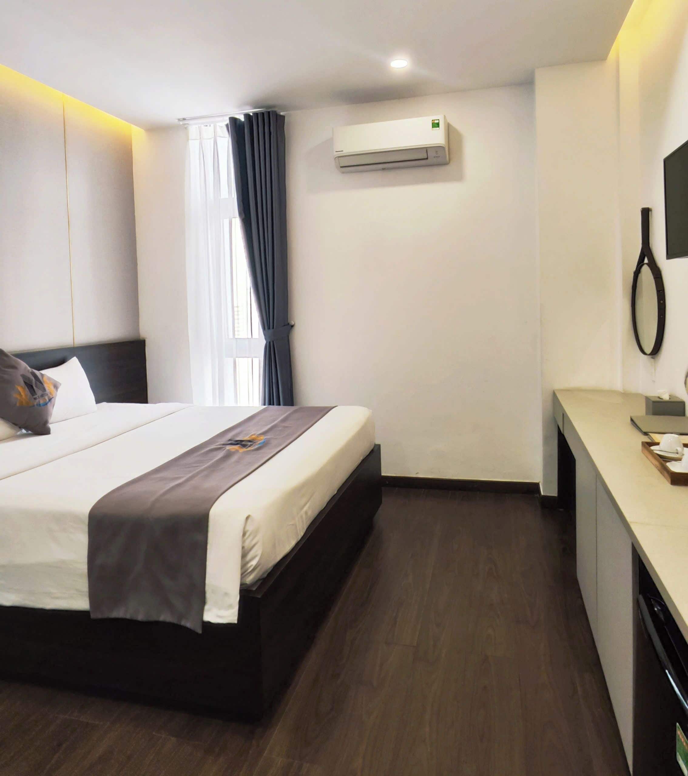 A hotel room with a bed, a desk, an air conditioning unit, and a window with curtains.