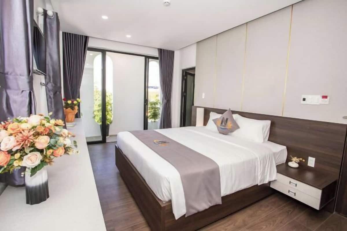 A modern bedroom with a large bed, bedside tables, a nightstand, a window with curtains, and a view of greenery outside.