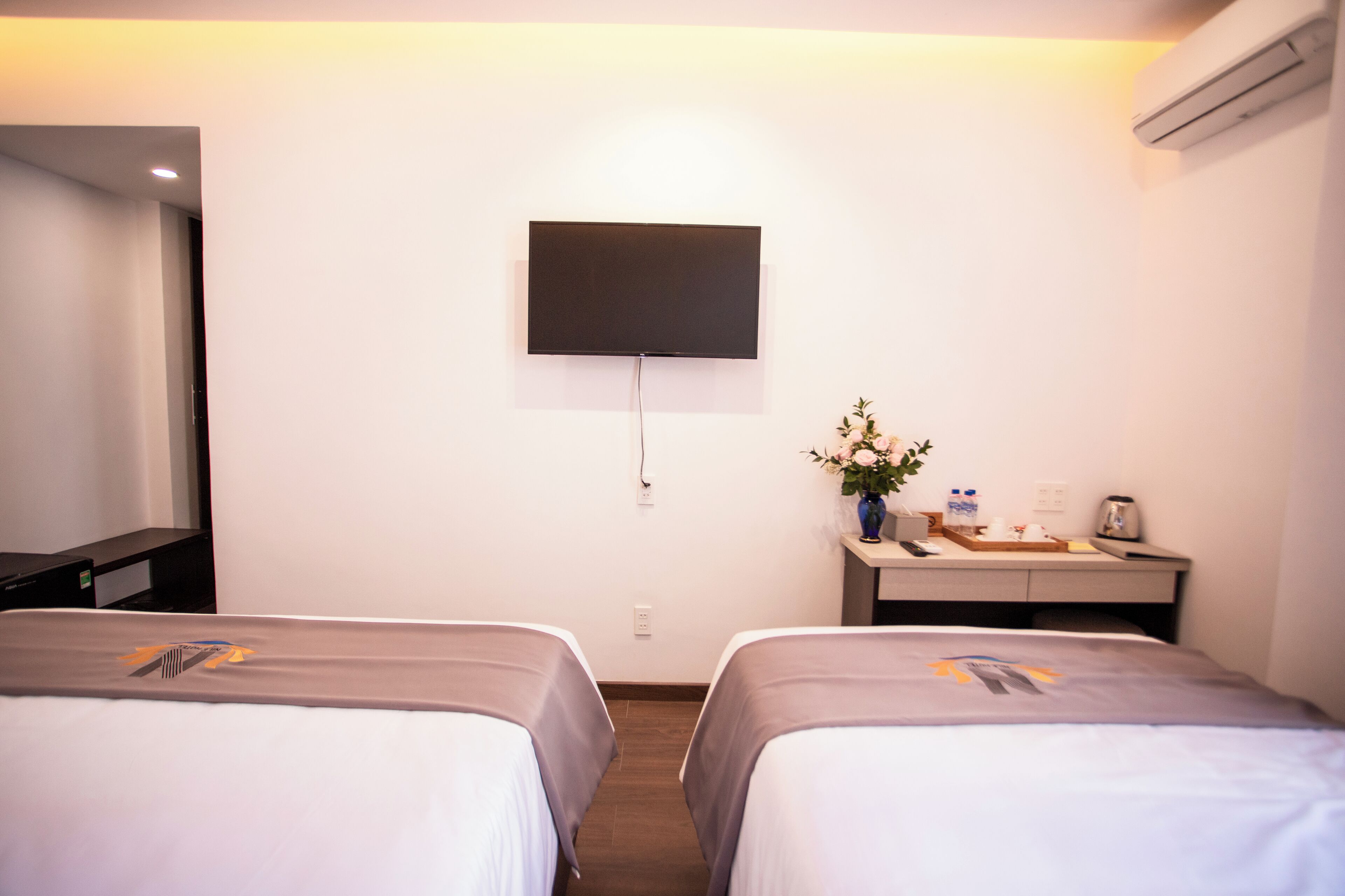 A hotel room with two beds, a flat-screen TV mounted on the wall, a desk with a vase of flowers, and an air conditioning unit.