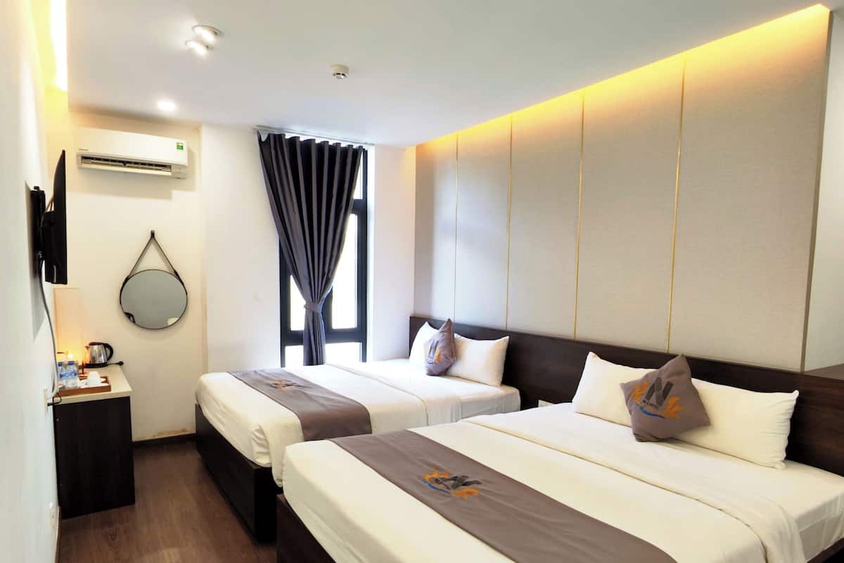 A hotel room with two beds, a bedside table with a lamp, a mirror, and an air conditioning unit.