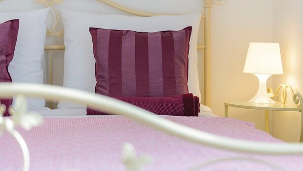 Ein ordentlich bezogenes Bett mit rosa Bettwäsche und einem weißen Kopfteil. Ein Nachttisch mit Lampe und einem kleinen dekorativen Gegenstand ist vorhanden.