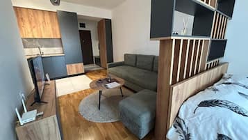 A modern, compact living space with a sofa, ottoman, and a bed with a patterned cover. There is a small round table with a rug, a TV, and a wooden cabinet with a flat-screen TV.