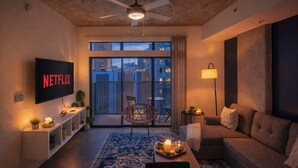 Una sala de estar moderna con un televisor de pantalla plana mostrando el logotipo de Netflix, un ventilador de techo, un balcón con vistas a la ciudad y una zona de estar acogedora con un sofá y una pequeña mesa.