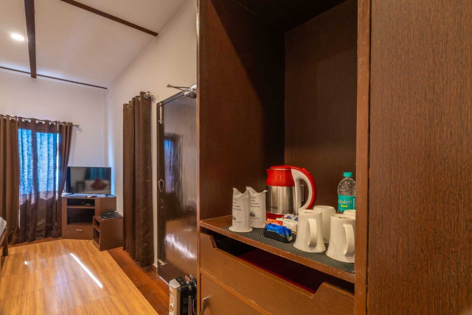 Habitación de hotel con piso de madera, un televisor sobre un mueble, cafetera, una botella de agua y una repisa con varios objetos.