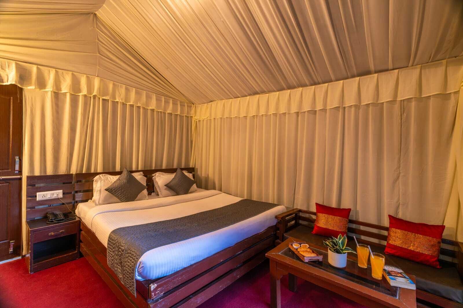 A hotel room with a large bed, a bedside table with a lamp, a small table with snacks and drinks, and a red carpet.