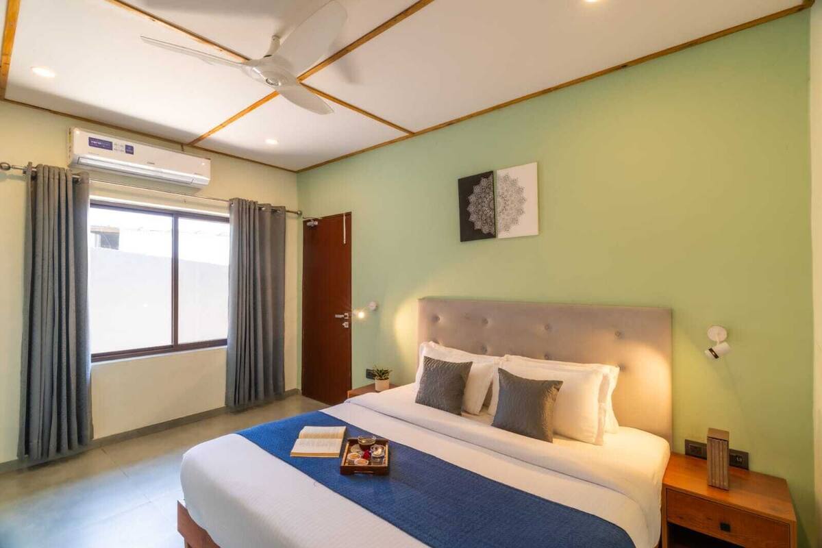 A hotel room with a bed, a ceiling fan, a window with curtains, and a wall-mounted artwork.