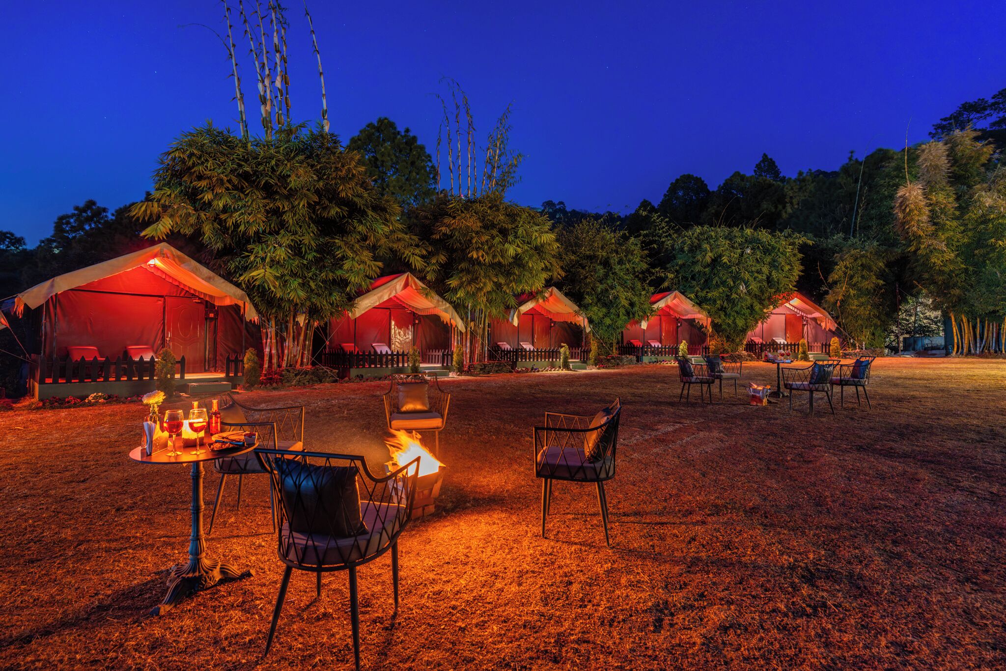 A series of red-roofed tents with tables and chairs arranged around a central fire, set in a grassy area with trees in the background.