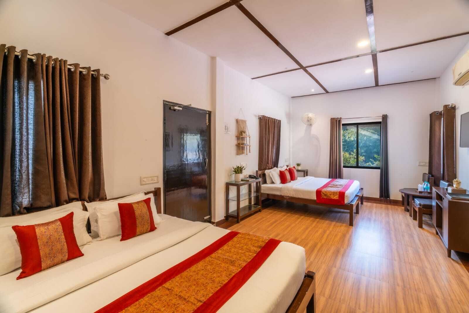 A hotel room with two beds, a desk, a chair, a small table, and a window with a view of greenery.