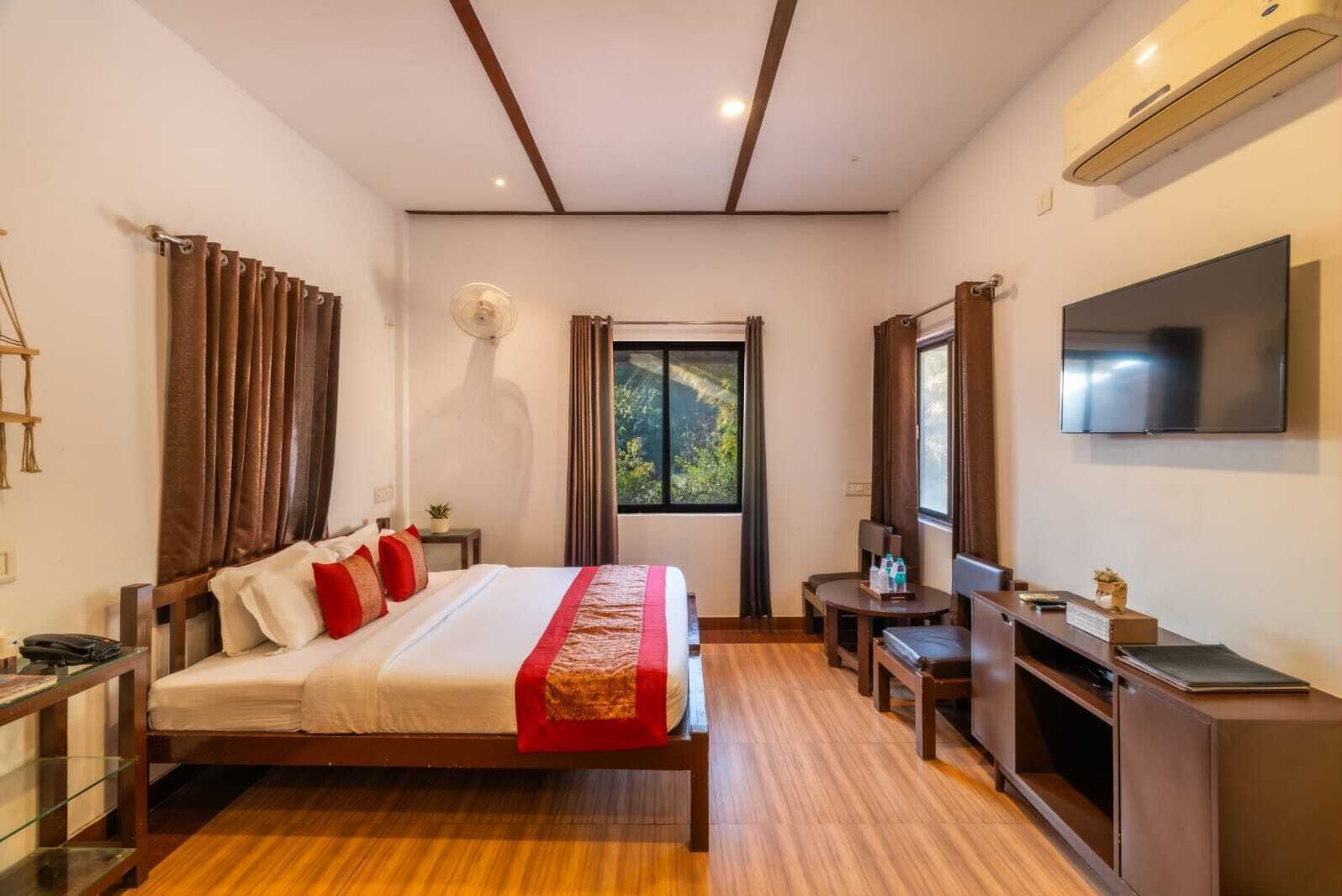 A modern hotel room with a wooden bed, a flat-screen TV, a desk, and a large window offering a view of greenery.