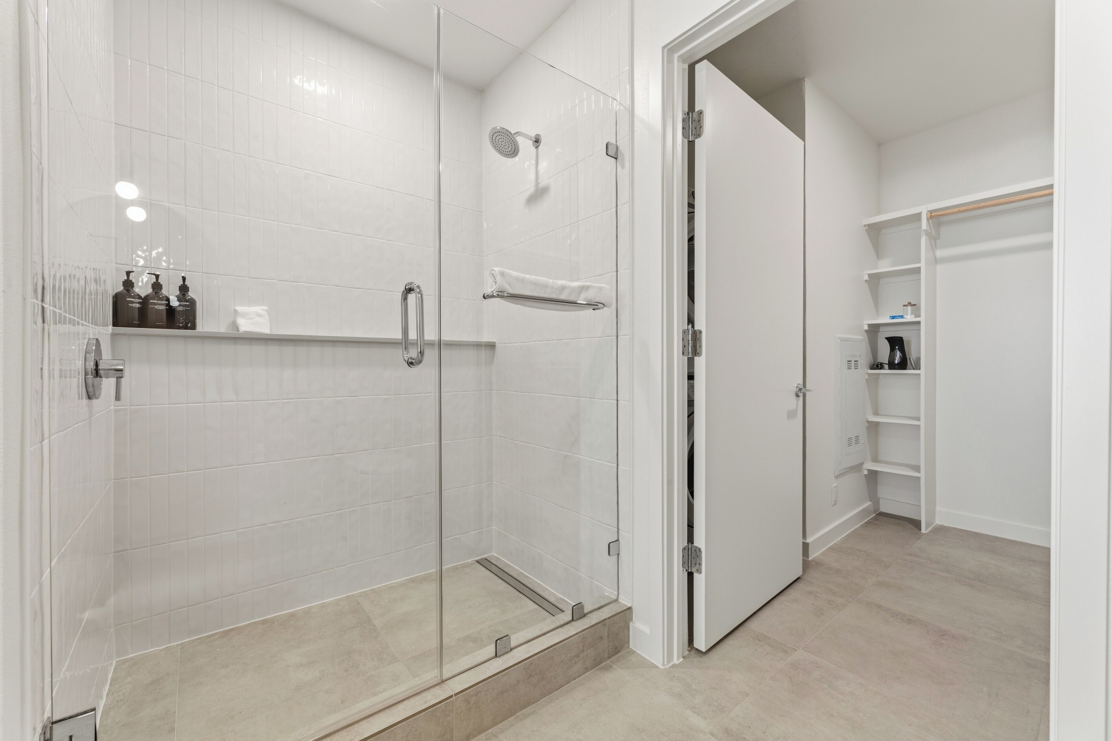 A modern bathroom with a glass-enclosed shower, a white tiled wall, and a walk-in closet with shelves.