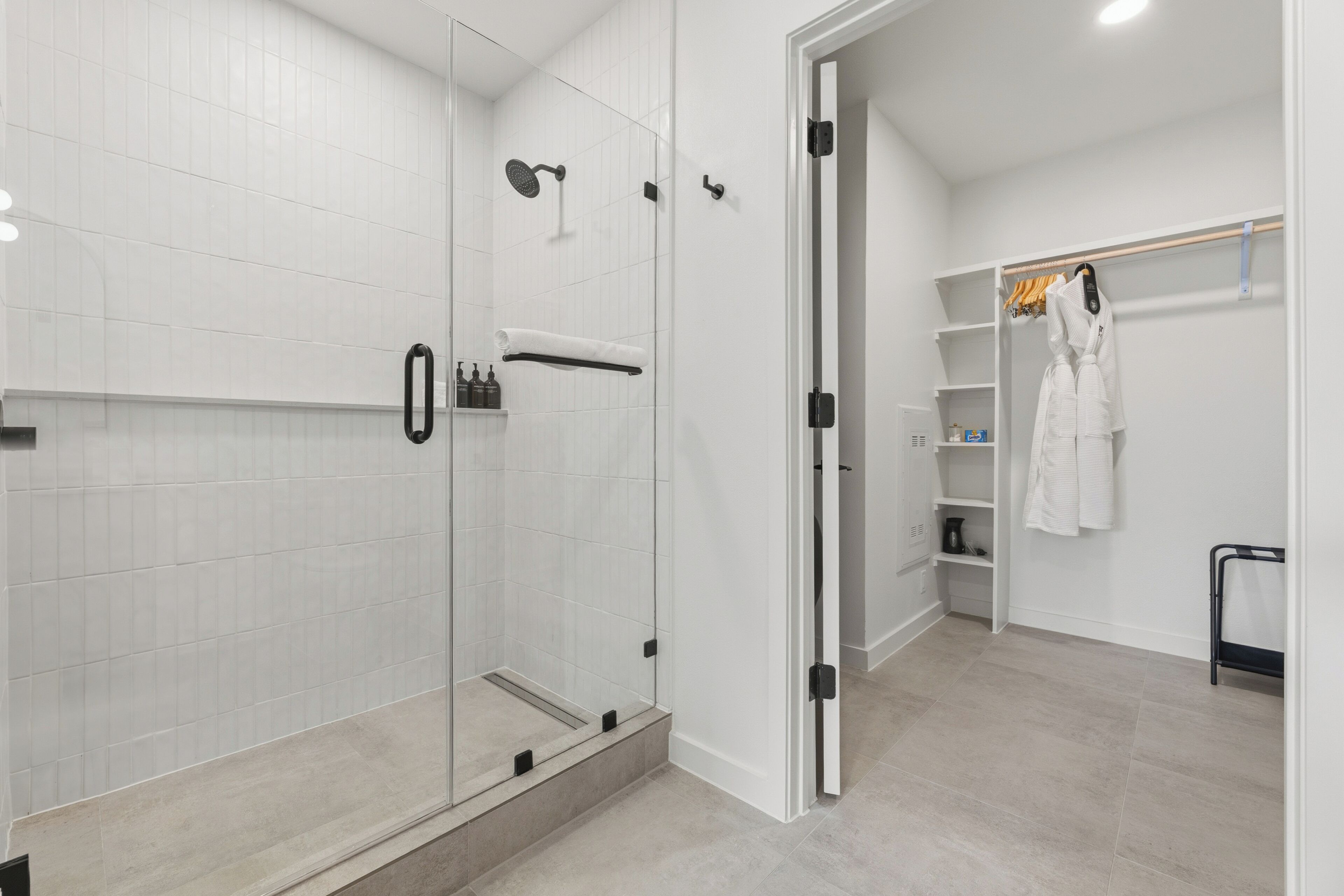 A modern bathroom with a glass-enclosed shower, a white tiled wall, and a walk-in closet with shelves and hanging robes.