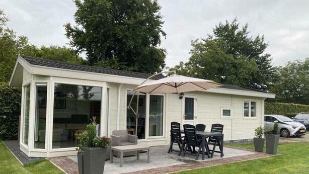 A modern single-story house with a patio area featuring a table and chairs, a large umbrella, and potted plants. - Minicamping De Lindenhoeve - Chalet Alpaca (Nistelrode)