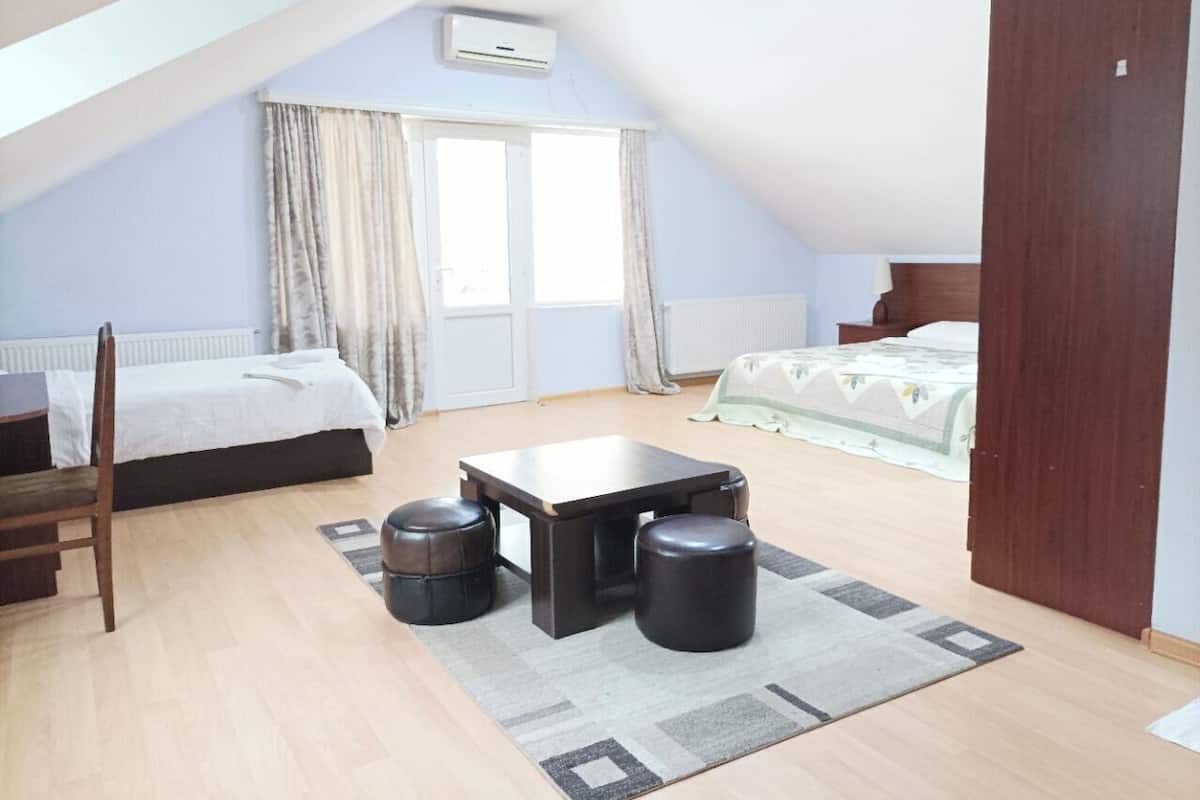 A bedroom with a bed, a small table, two stools, a chair, and a window with curtains.