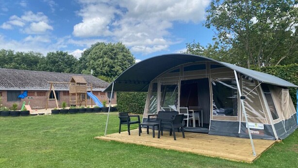 Un terrain de camping avec une grande tente, une terrasse en bois et un bâtiment en briques, avec un toboggan bleu en arrière-plan.