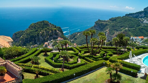 Une villa située sur la côte, avec un toit recouvert de tuiles en terre cuite, une piscine et un jardin bien entretenu agrémenté de haies et de palmiers.