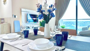 A modern dining area with a table set for four, a vase of flowers, and a city view through large windows. - Macrolink Medini Legoland by Stayrene (Iskandar Puteri)