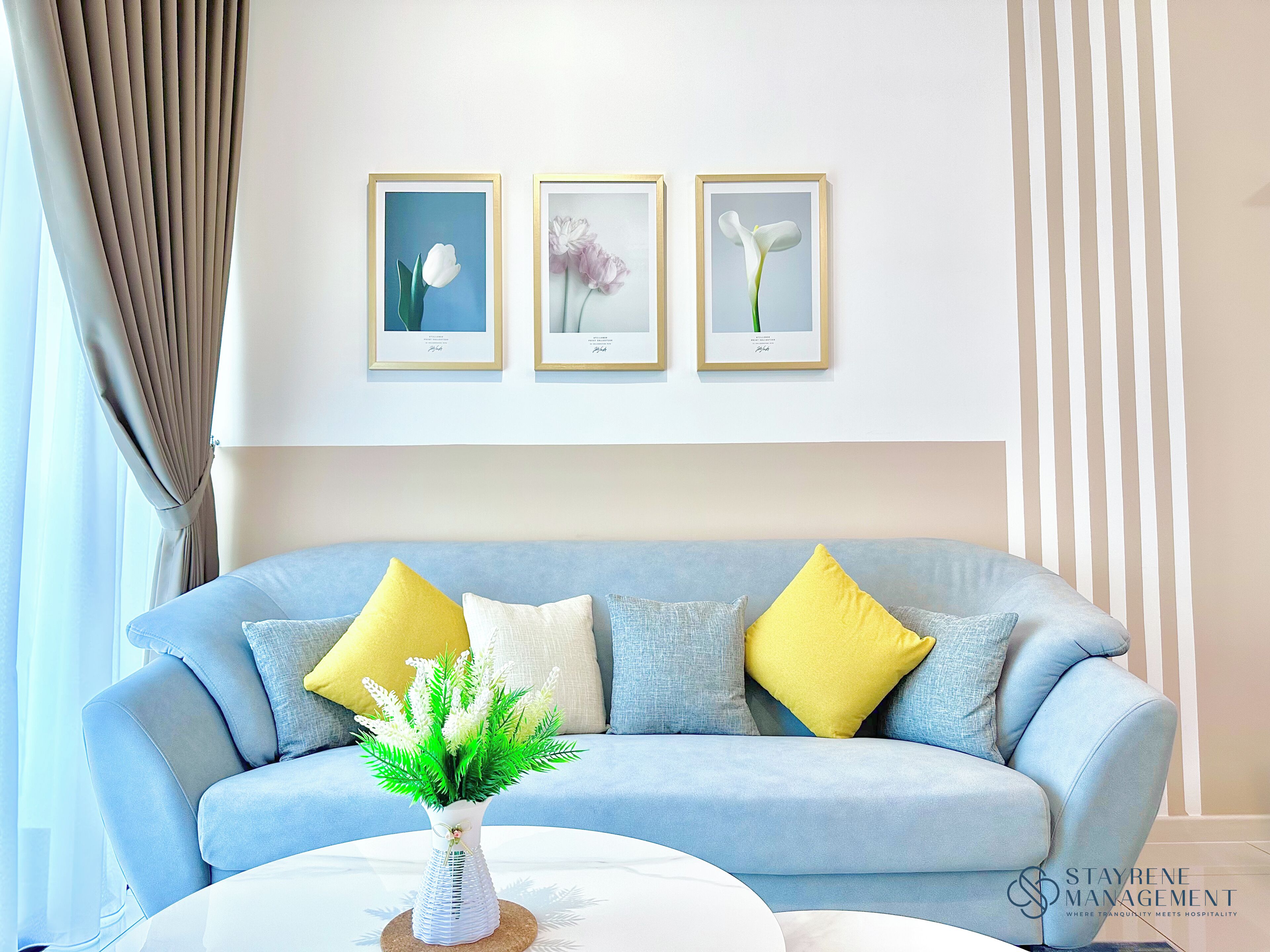 A living room with a light blue sofa, yellow and white cushions, a white vase with green foliage, and three framed floral paintings on the wall.