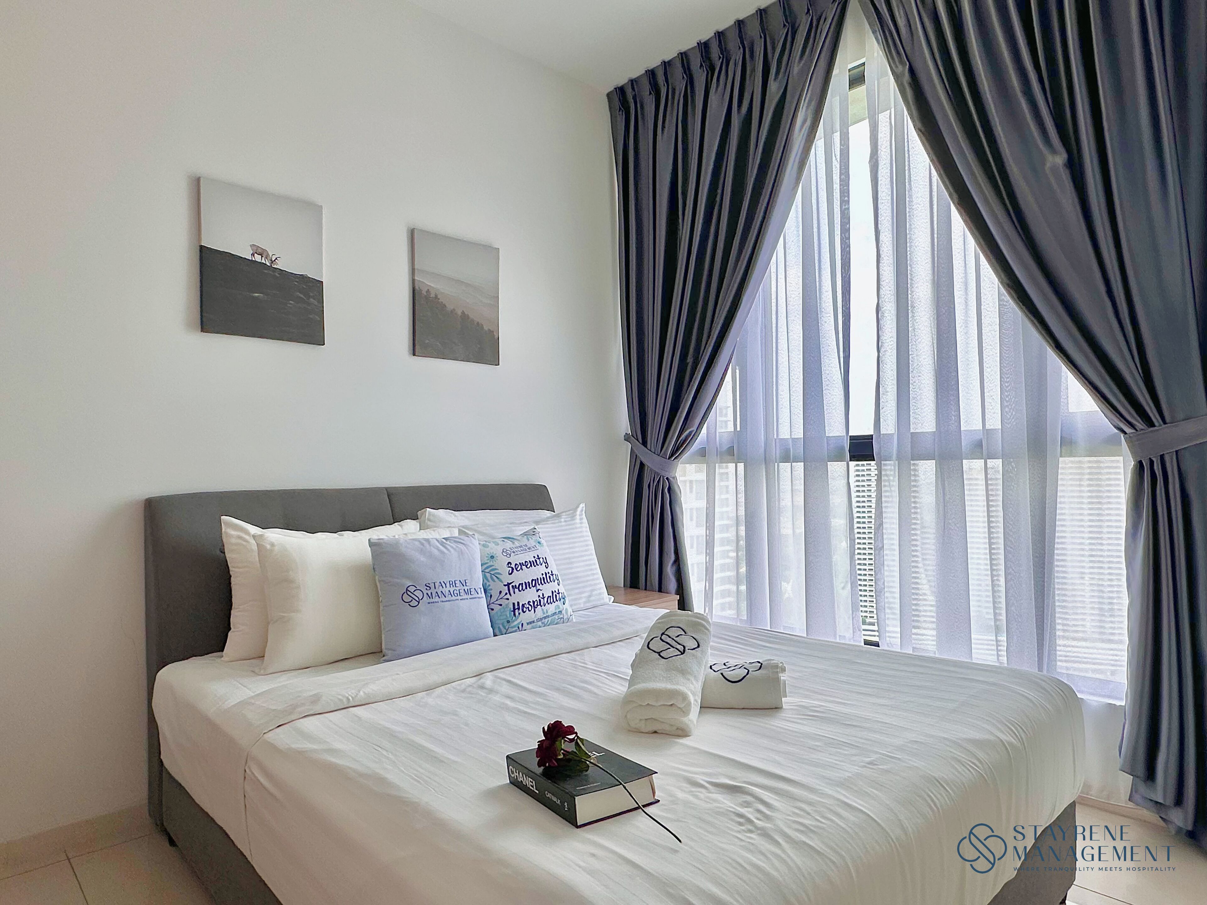 Una cama bien hecha con sábanas blancas, dos almohadas, un libro y una rosa.