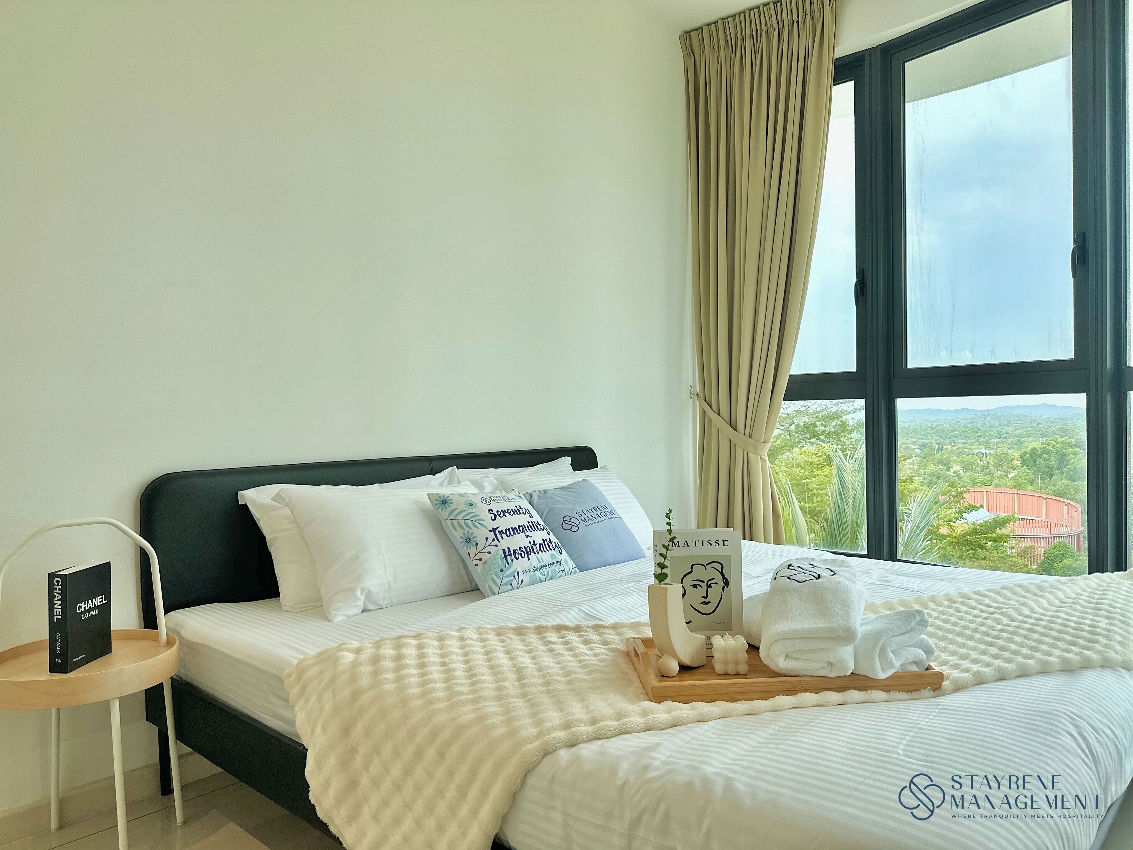 Habitación de hotel con cama, almohadas, un atril con artículos de aseo, una caja de Chanel y vistas al exterior con follaje.