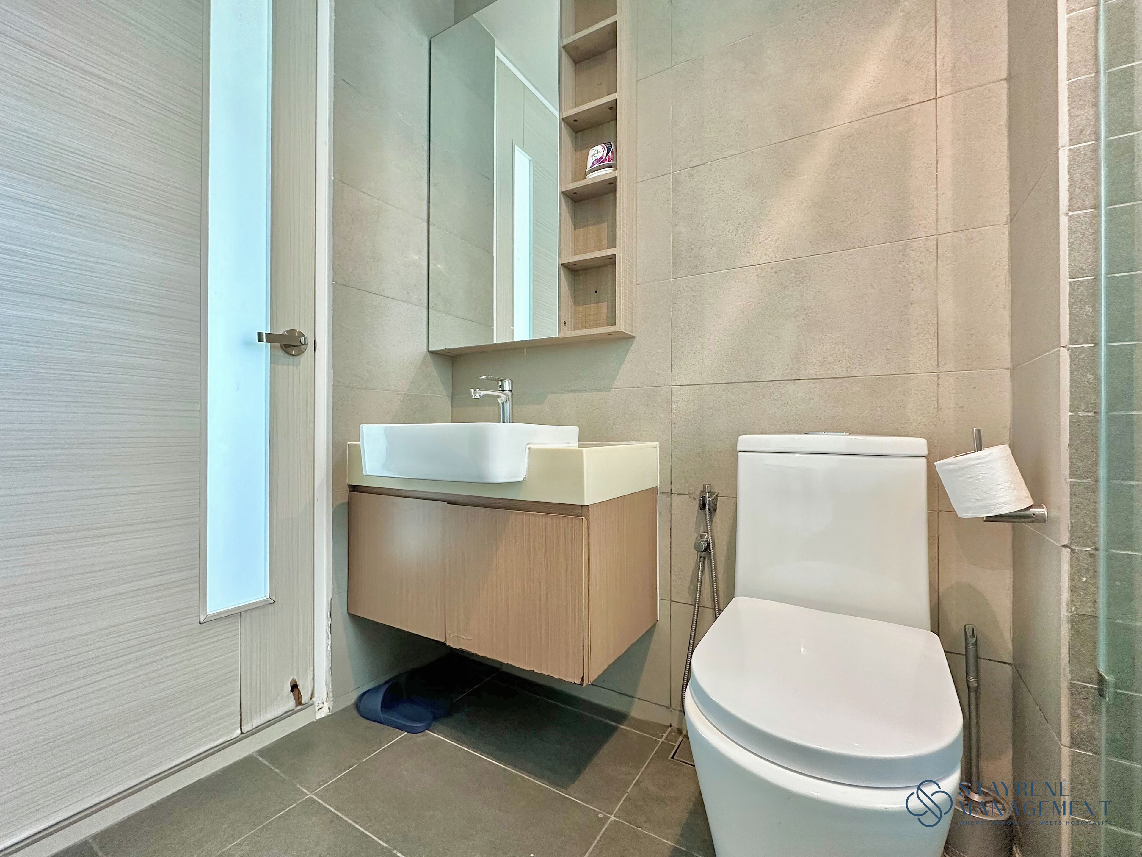 Un baño moderno con lavamanos blanco, tocador de madera, inodoro y cabina de ducha con mampara de vidrio.