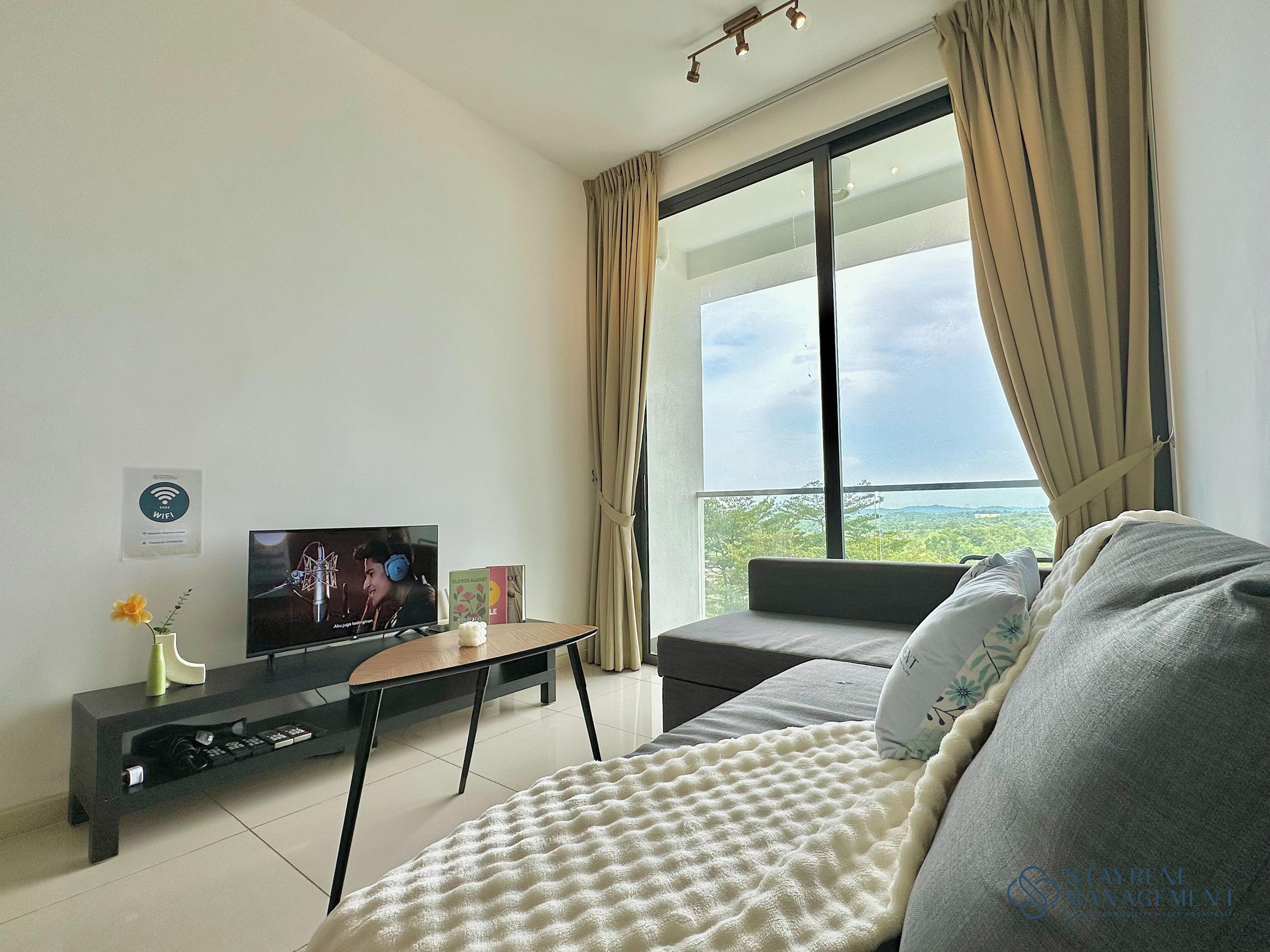 A modern living room with a flat-screen TV, a sofa, a coffee table, and a large window offering a view of the outdoors.