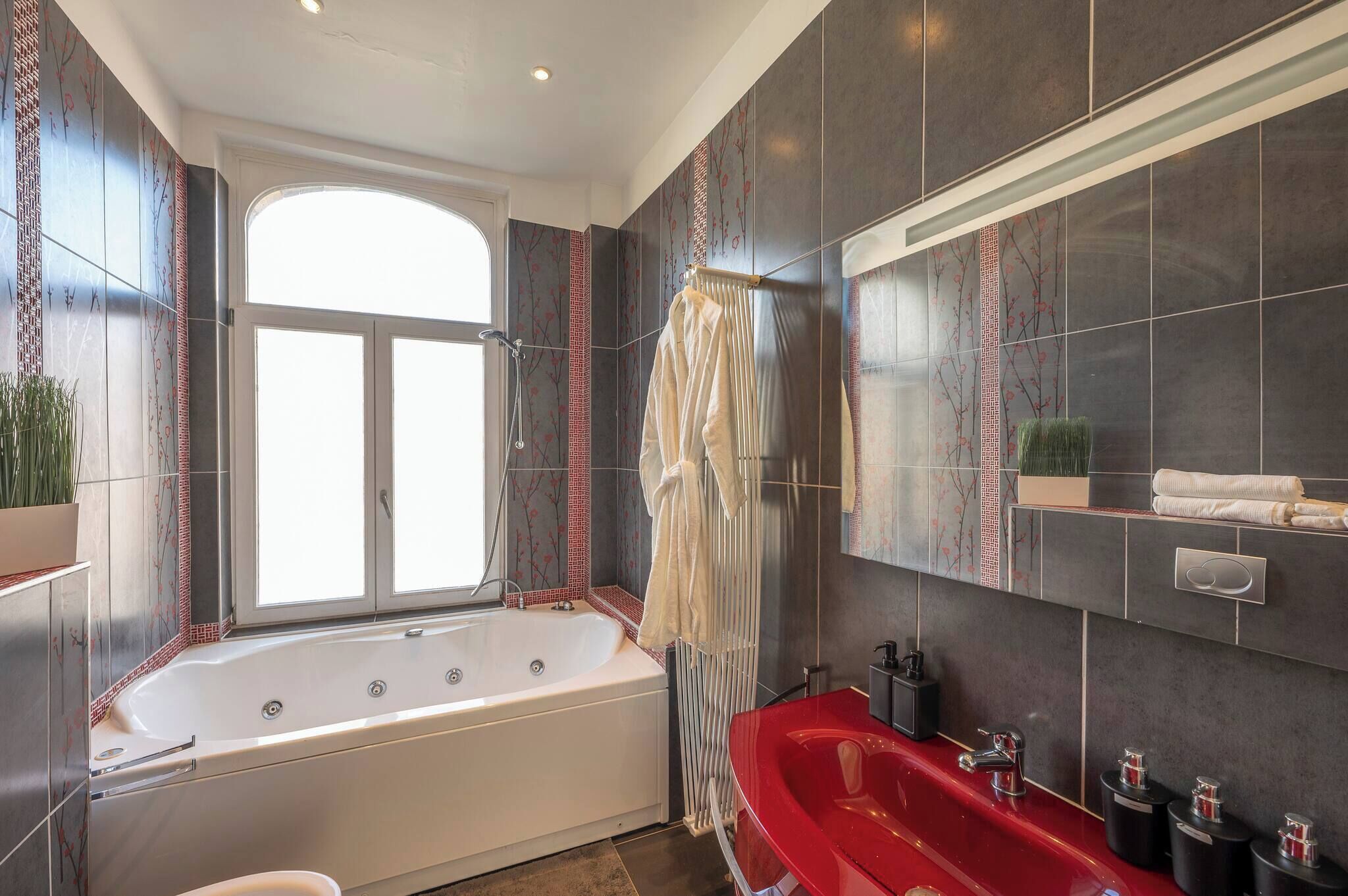 A modern bathroom with a large bathtub, a red sink, and a window providing natural light.