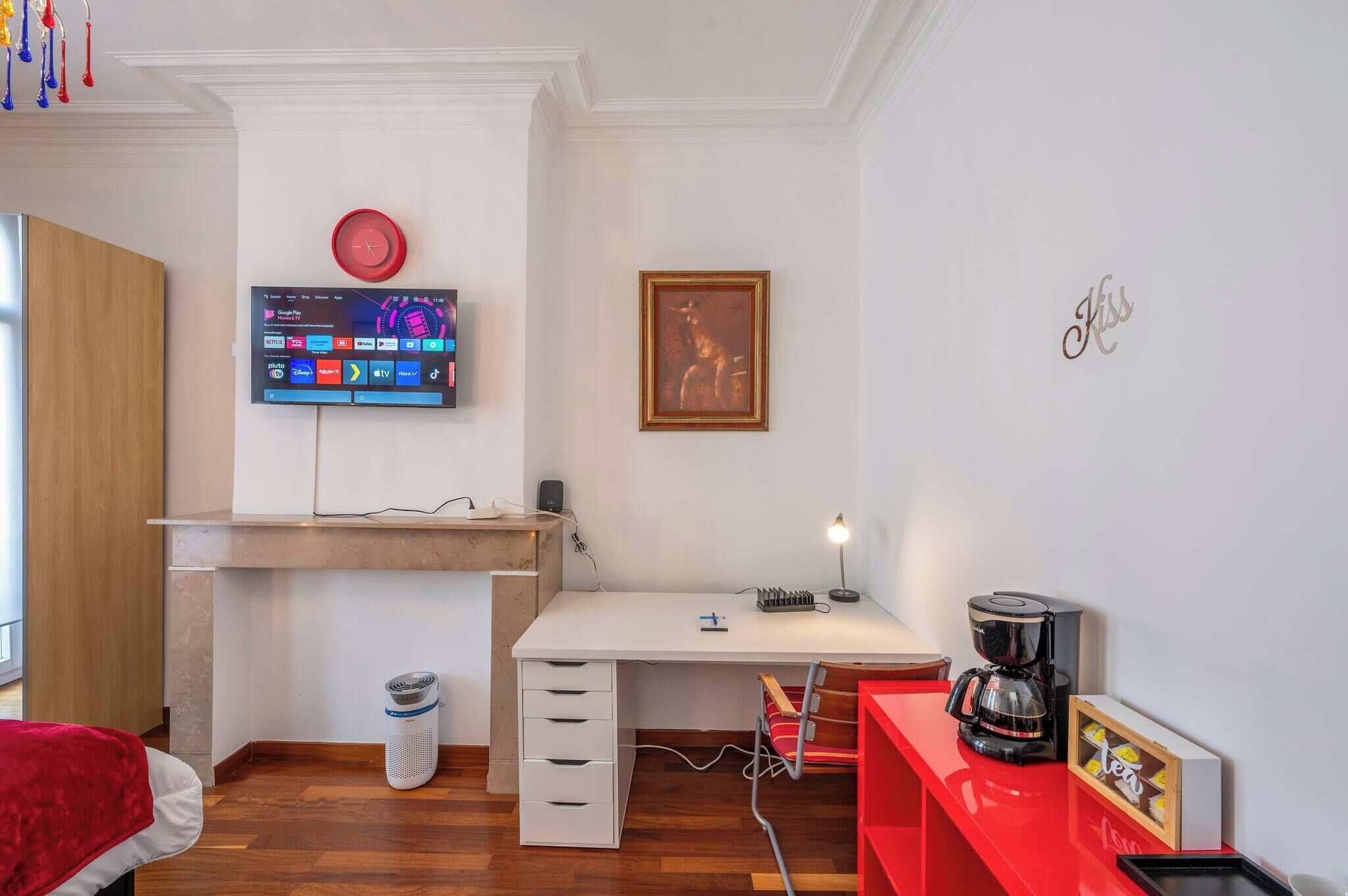 Een moderne kamer met een bureau, een wandtelevisie, een lijst met een afbeelding en een rode koffiezetapparaat.