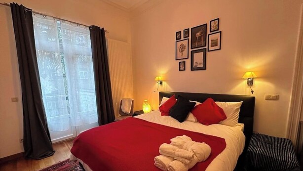 A hotel room with a large bed, two bedside lamps, a window with curtains, and a wall with framed pictures.