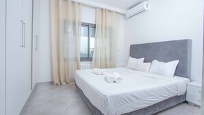 Ein modernes Schlafzimmer mit einem großen Bett, weißen Leinen, einem grauen Kopfteil und einem Fenster mit durchscheinenden Vorhängen.