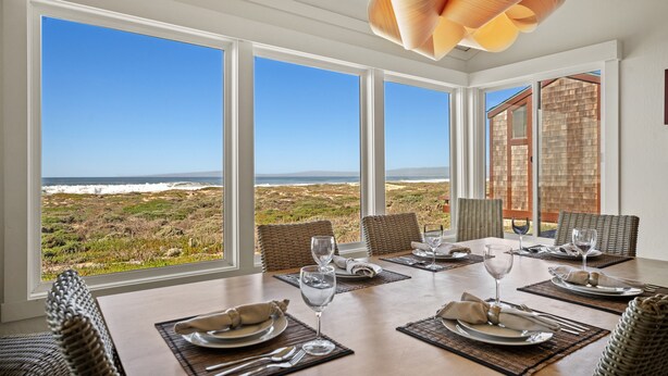 Spiseområde med et bord dækket til fire personer, store vinduer med udsigt over havet og stranden, samt en moderne loftlampe.