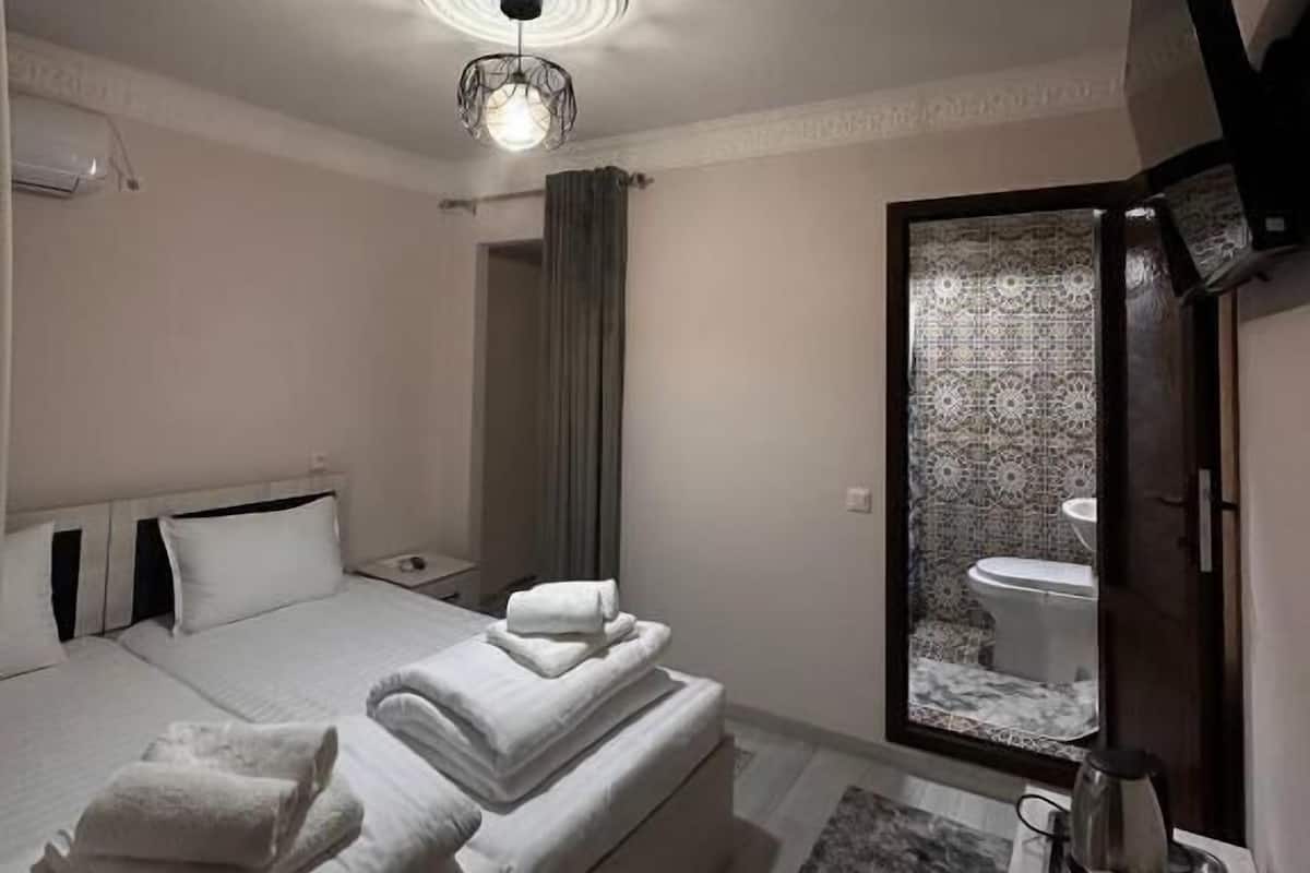 A modern hotel room with a bed, a bedside table with a coffee maker, a TV mounted on the wall, and a bathroom visible through an open door.