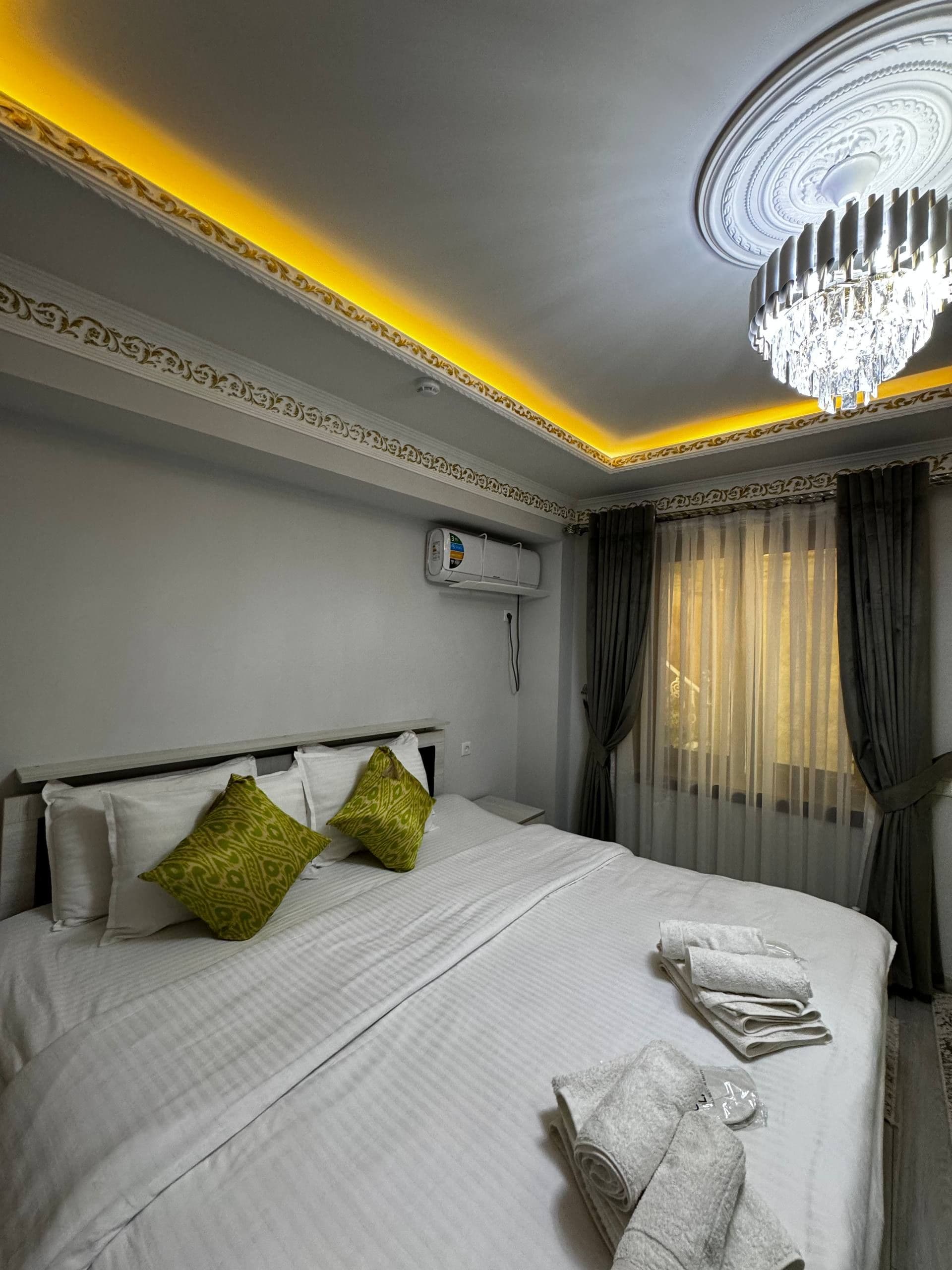 A hotel room with a large bed, a chandelier, an air conditioning unit, and a window with curtains.