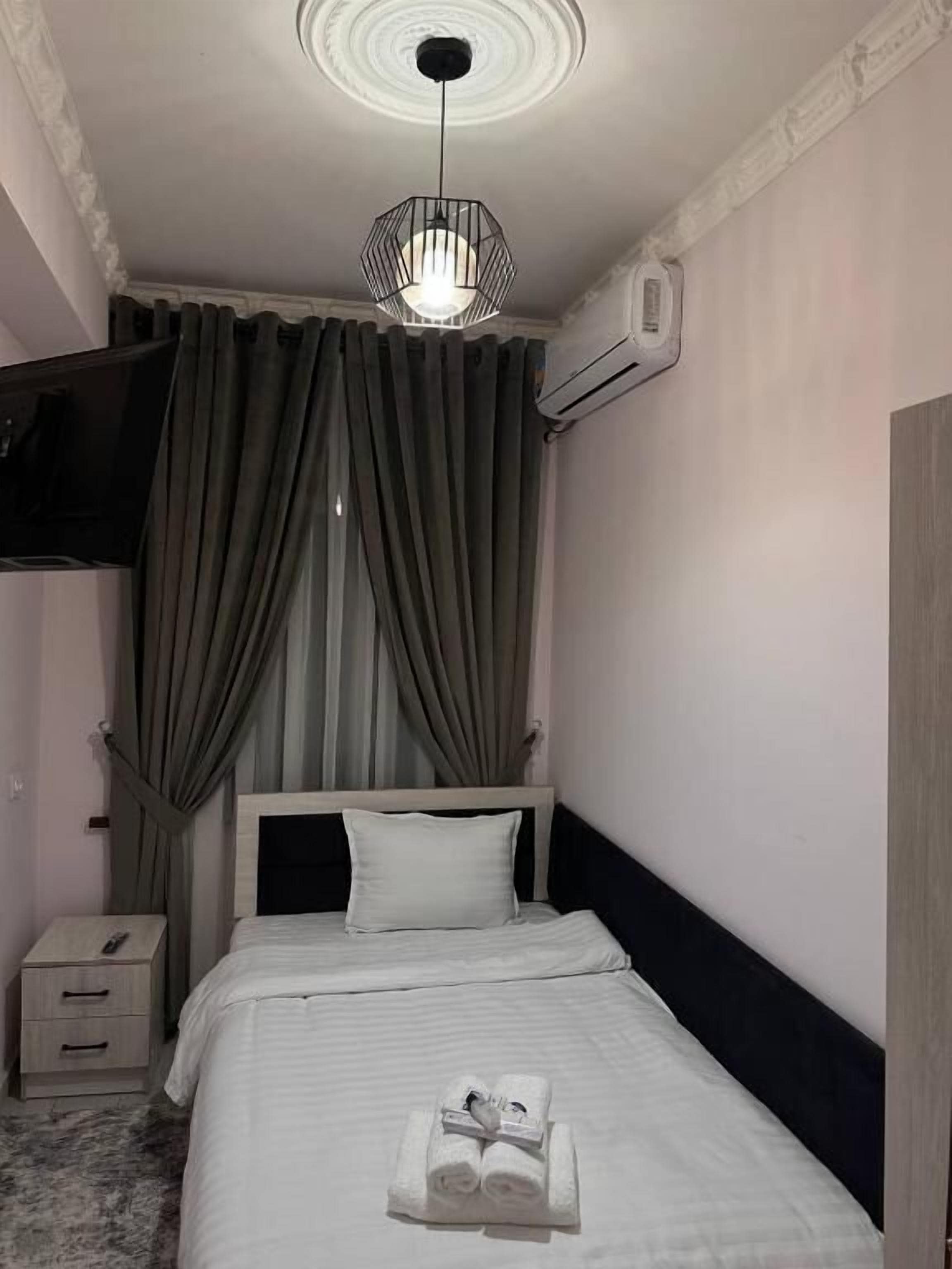 A neatly made bed with a white comforter and a folded towel, a bedside table with a lamp, and a ceiling light fixture.