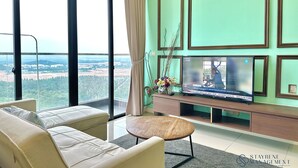 A modern living room with a large TV, comfortable seating, and a view of a green landscape through large windows. - D'Pristine Medini by Stayrene (Iskandar Puteri)