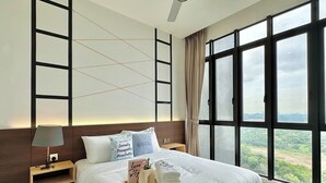 A modern hotel room with a large bed, a bedside table with a lamp, a tray with towels and a book, and a view of greenery through large windows. - D'Pristine Medini by Stayrene (Iskandar Puteri)