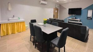 Habitación de hotel moderna con zona de comedor, sofá, televisor y baño con lavabo y espejo.